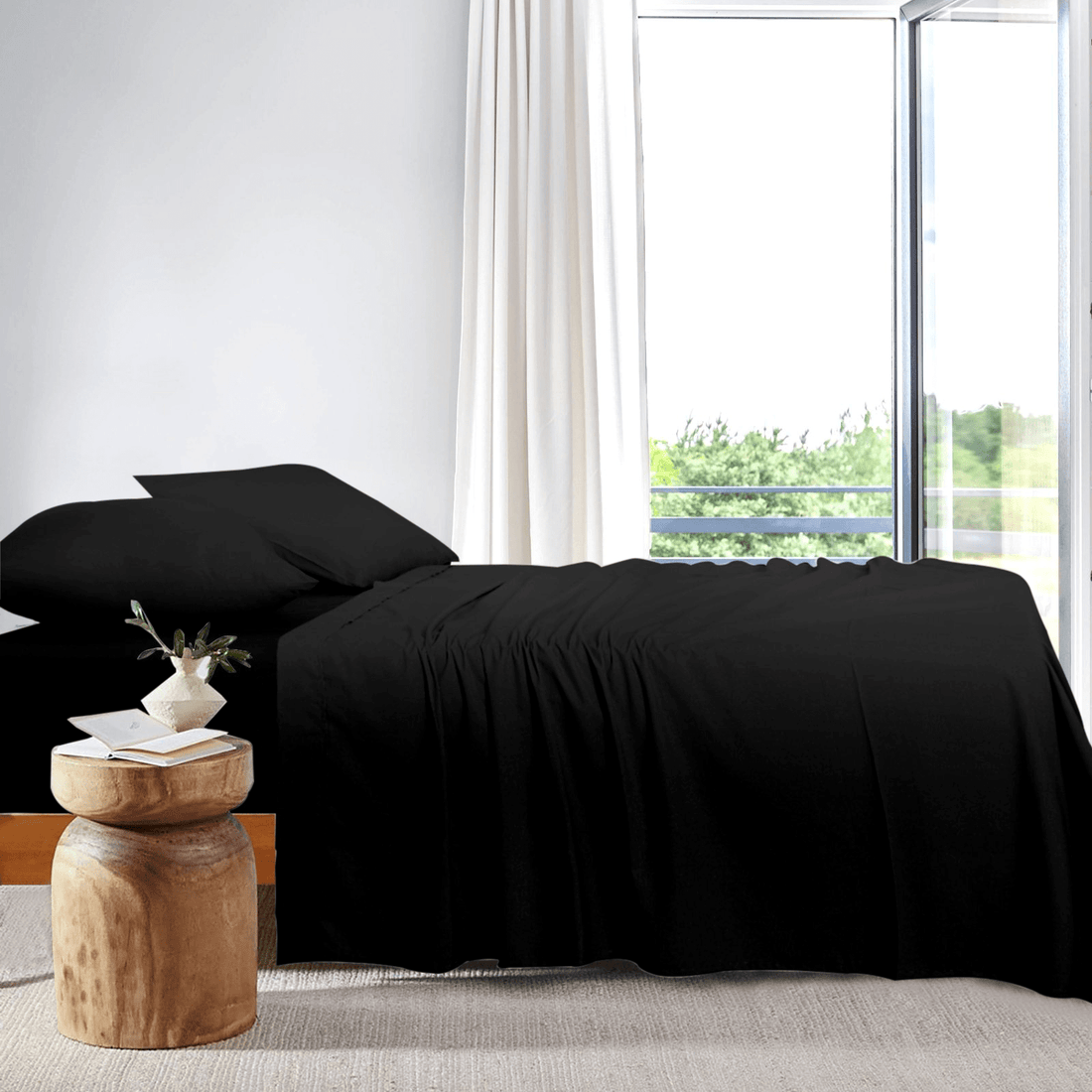 Side view of a neatly made bed dressed in black bamboo sheets next to a large window in a bright bedroom