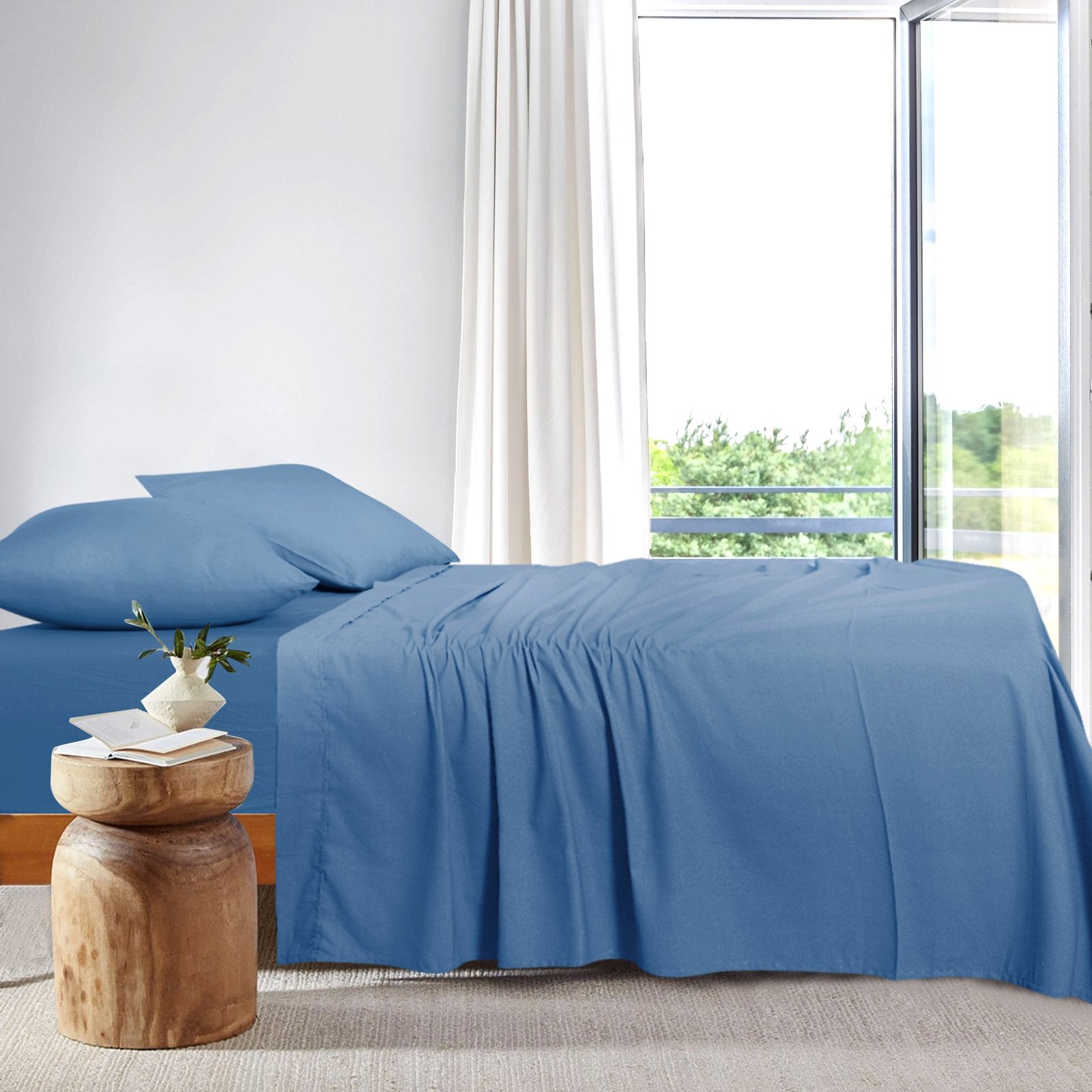 Side view of a modern bed neatly dressed in blue bamboo sheets positioned next to a bright window.