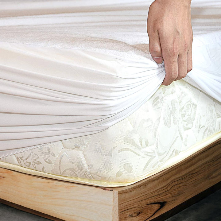 Hand pulling back a white terry toweling waterproof mattress protector to reveal the fitted mattress corner beneath