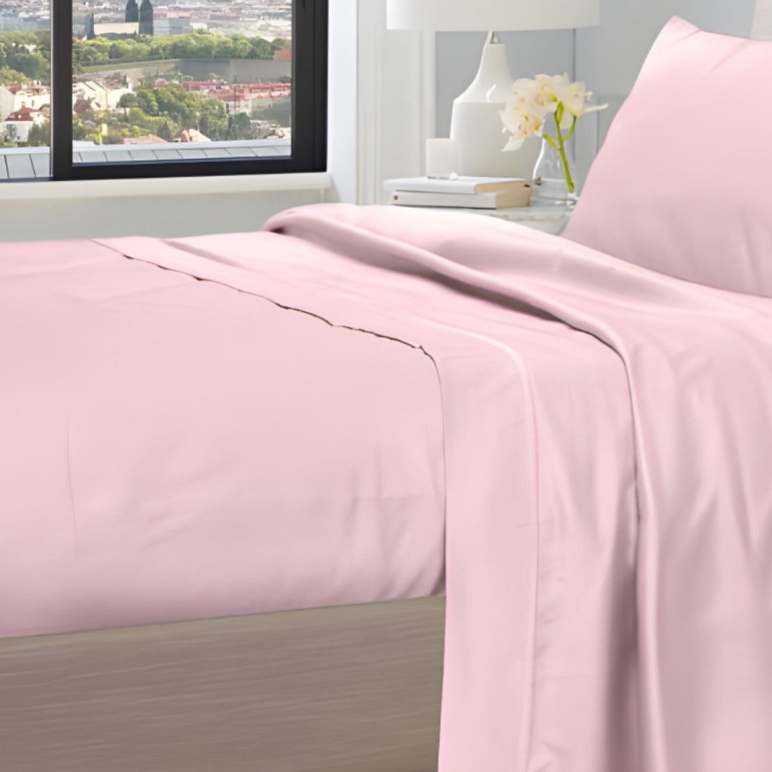 Baby pink bed sheets draped over a bed near a window with city view