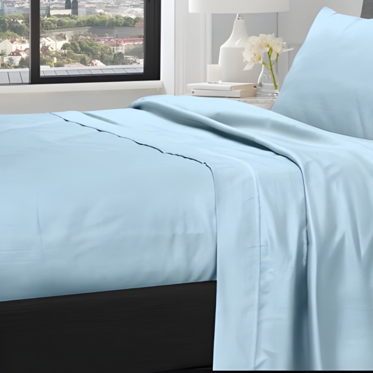 Light blue bed sheets draped over a bed near a window with city view